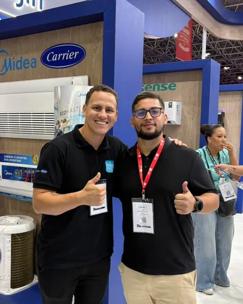 Dois profissionais sorrindo e fazendo sinal de positivo em uma feira do setor de climatização, diante de estandes de fabricantes de ar-condicionado
